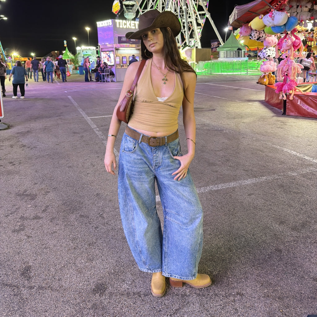 A woman stands at a nighttime fairground wearing a tan cropped tank top, wide-leg light blue jeans, tan boots, and a brown cowboy hat, with a brown shoulder bag. A Ferris wheel and carnival games are visible in the background.
