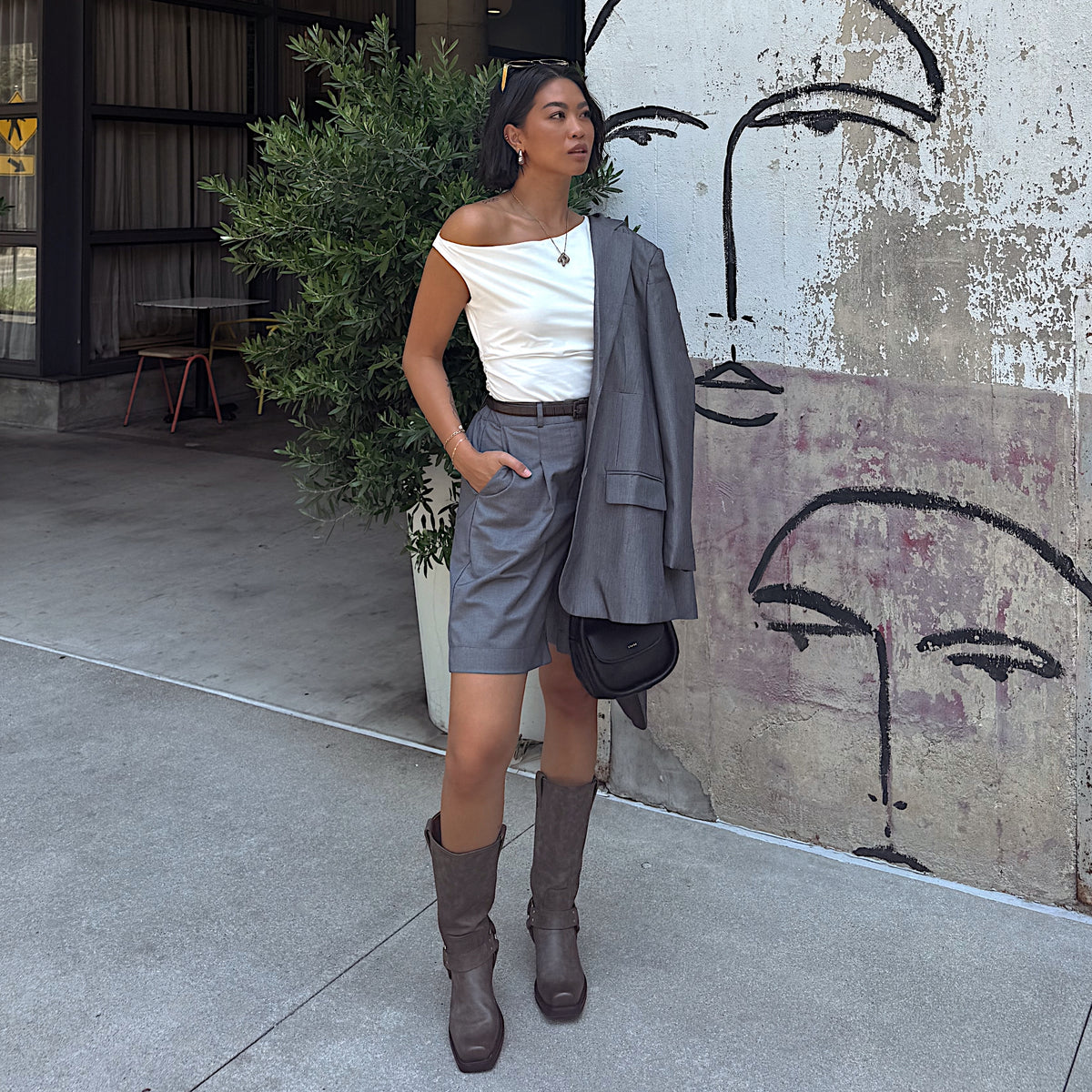Woman wearing gray knee-high boots with buckle details, gray tailored shorts, a white off-the-shoulder top, and a gray blazer while standing outdoors.