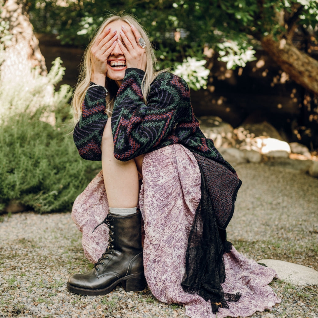 A woman crouching outdoors on a gravel path, covering her face with her hands. She is wearing a dark green and burgundy patterned knit sweater, a dusty pink patterned maxi skirt, and black lace-up combat boots.