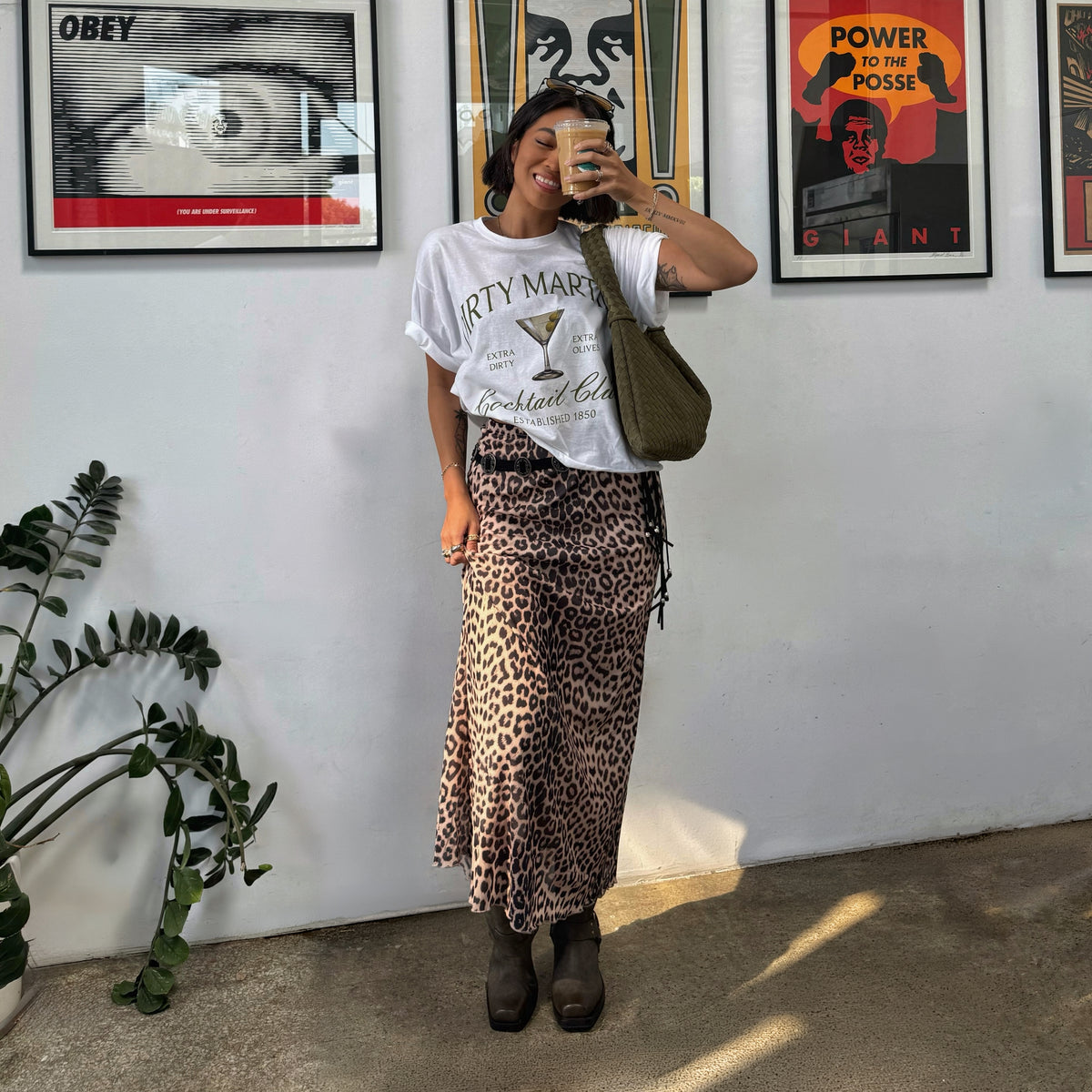 Person wearing dark brown ankle boots with a leopard-print midi skirt and graphic T-shirt, holding a coffee cup and standing indoors beside framed posters.