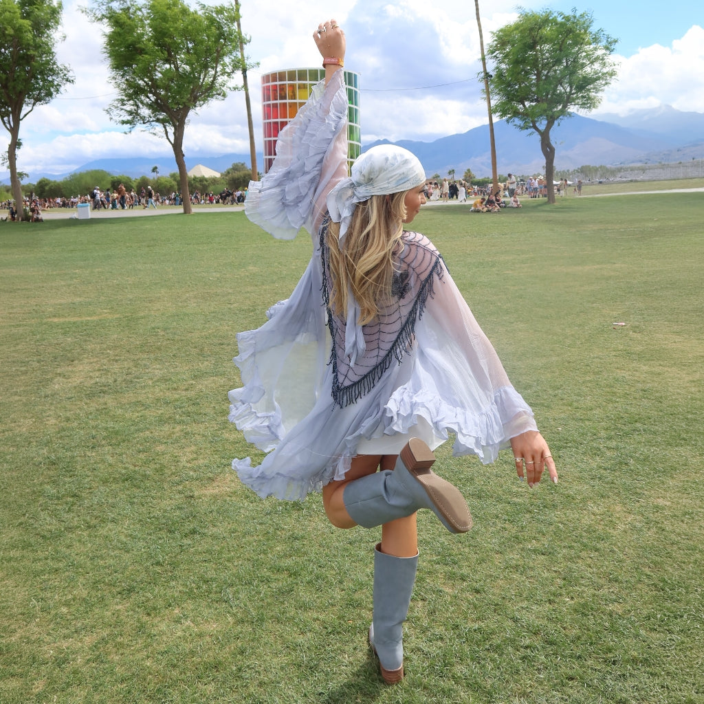 A woman outdoors on a grassy field wearing a sheer light blue ruffled kimono-style cover-up over a white top, with gray knee-high boots and a white headscarf. Trees, mountains, and distant people are visible in the background.
