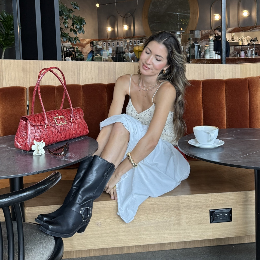 A woman sits on a wooden bench in a cafe setting, wearing a white lace-trimmed spaghetti-strap dress, black leather boots, and gold jewelry. A bright red textured handbag rests on the table next to her.