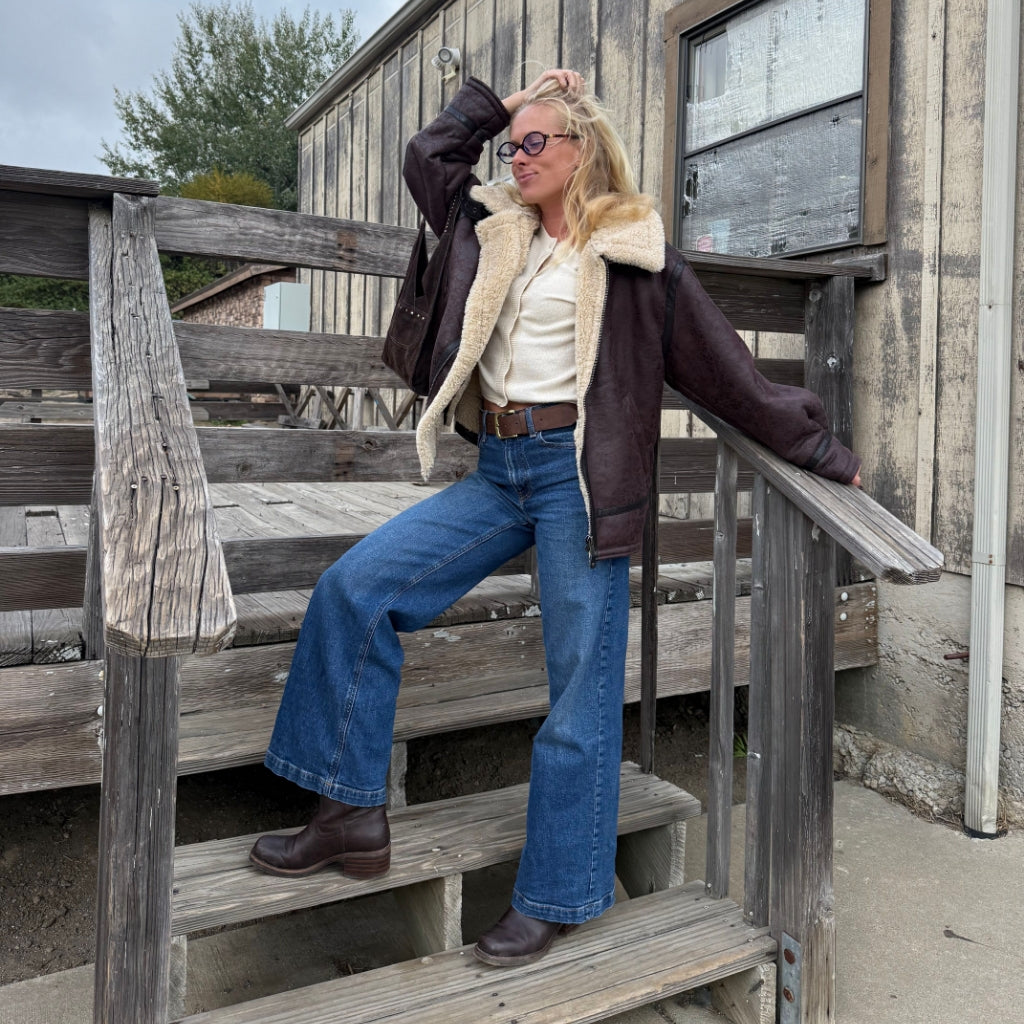 A woman wearing a dark brown shearling-lined aviator jacket, blue wide-leg jeans, and brown boots poses on rustic wooden outdoor stairs in front of a weathered wooden building.