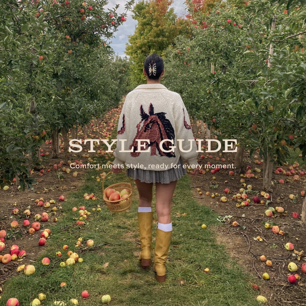 A person stands in the center of an apple orchard, viewed from behind, wearing a cream knit sweater with a large horse motif, a pleated gray skirt, yellow knee-high boots, and a black cap. They are holding a basket of apples.