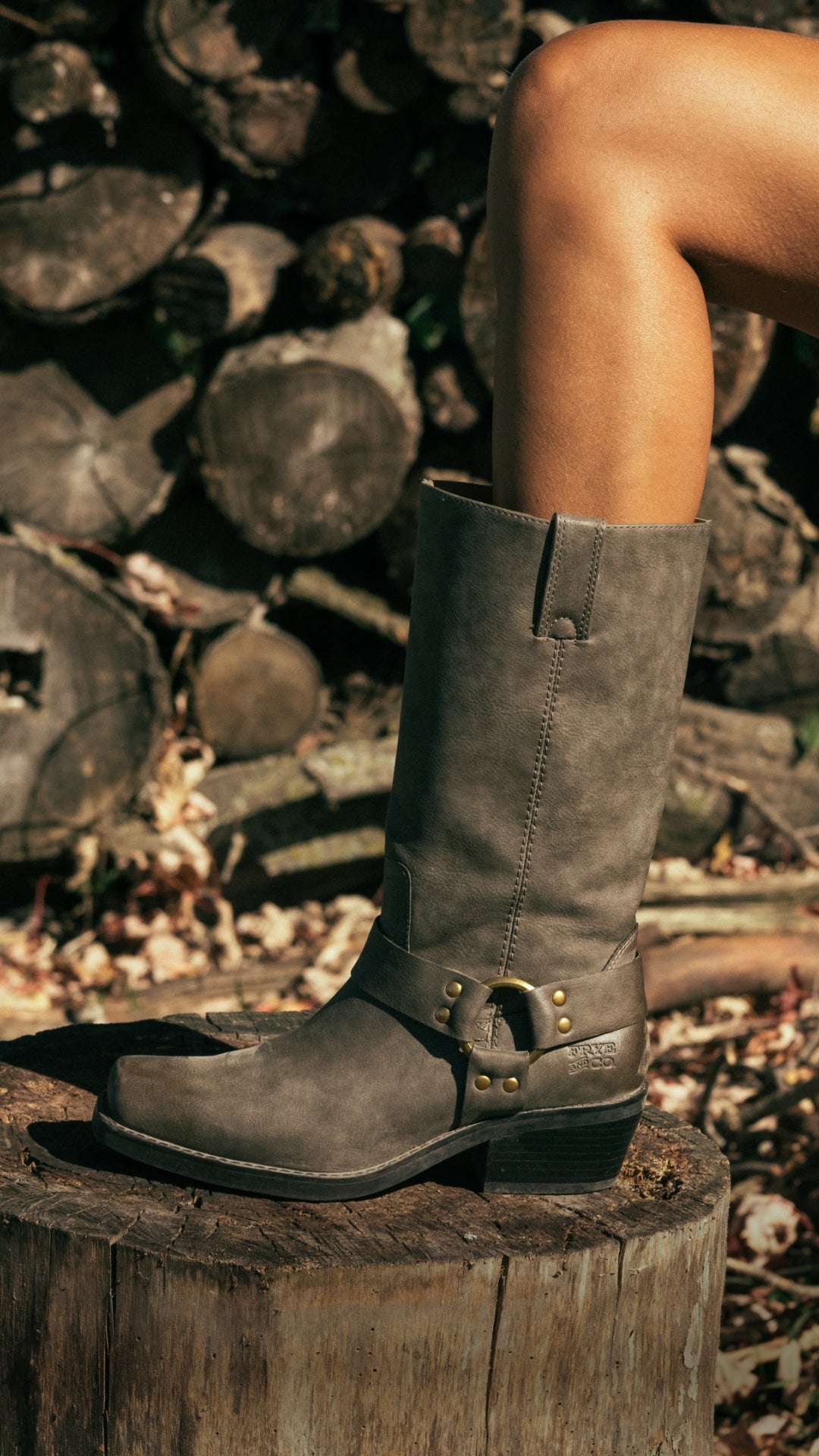 Close-up of a person wearing a gray leather knee-high boot with a decorative strap, brass buckle and studs, standing on a wooden tree stump outdoors.