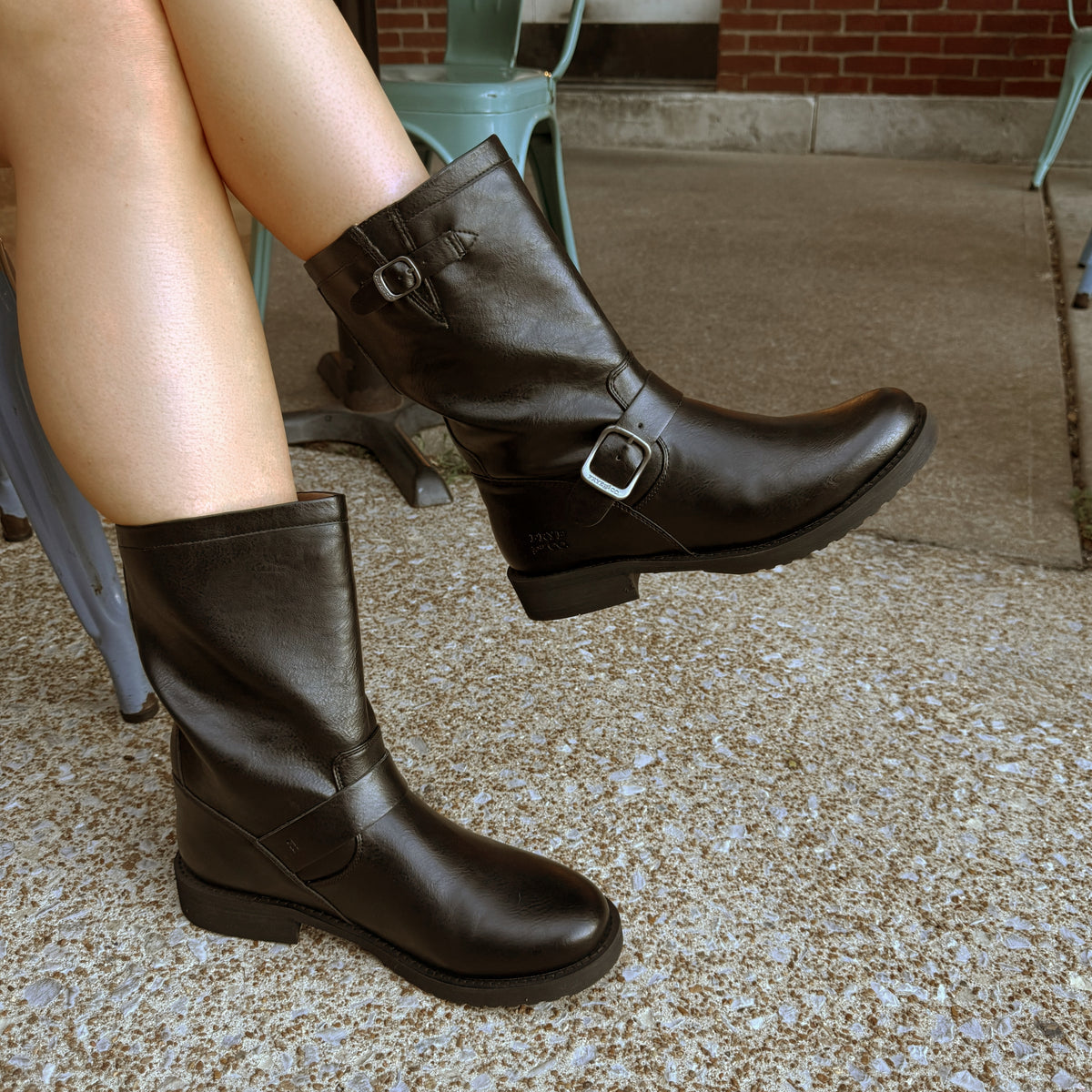 Close-up of a person wearing dark brown leather mid-calf boots with adjustable buckle straps and low heels, seated outdoors.