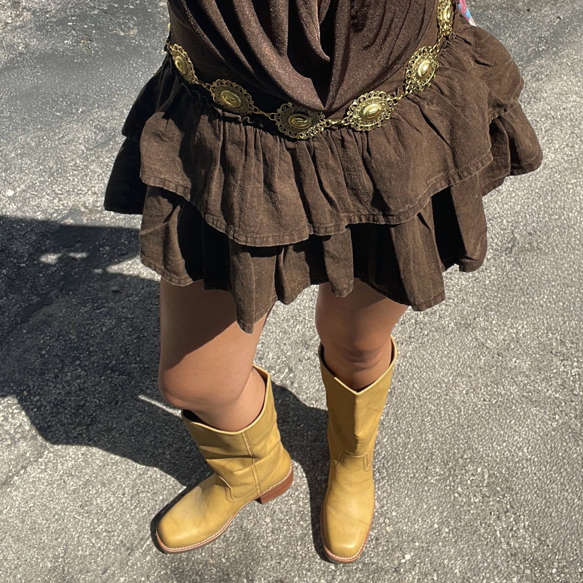 Close-up of a person wearing tan knee-high leather boots with low stacked heels, paired with a layered brown skirt and decorative gold belt.